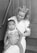 A woman in a nurse's uniform is holding a baby on her lap. The woman has curly hair and is smiling gently at the baby. The baby is wearing a sleeveless outfit and is looking slightly off to the side with a curious expression. They are sitting on a set of wooden steps, creating a simple, intimate scene.