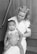 A woman in a nurse's uniform is holding a baby on her lap. The woman has curly hair and is smiling gently at the baby. The baby is wearing a sleeveless outfit and is looking slightly off to the side with a curious expression. They are sitting on a set of wooden steps, creating a simple, intimate scene.