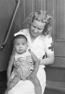 A woman in a nurse's uniform is holding a baby on her lap. The woman has curly hair and is smiling gently at the baby. The baby is wearing a sleeveless outfit and is looking slightly off to the side with a curious expression. They are sitting on a set of wooden steps, creating a simple, intimate scene.