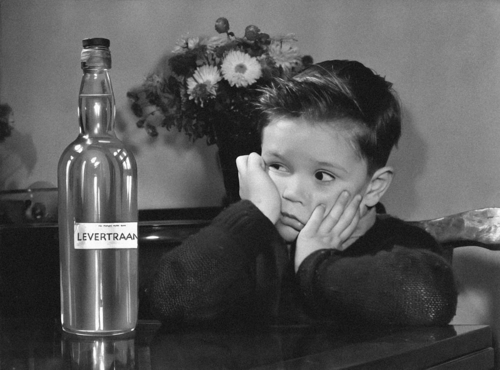 a black and white photo of a young boy sitting at a table with a bottle