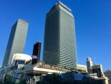 Two tall, modern skyscrapers dominate the scene, surrounded by palm trees and a bright blue sky. The buildings have a sleek, glass exterior with the name 'Cosmopolitan' visible on one of them. Below, there are various structures, including a billboard and what appears to be other parts of a commercial complex.