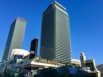 Two tall, modern skyscrapers dominate the scene, surrounded by palm trees and a bright blue sky. The buildings have a sleek, glass exterior with the name 'Cosmopolitan' visible on one of them. Below, there are various structures, including a billboard and what appears to be other parts of a commercial complex.