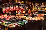 A vibrant market stall filled with a variety of fresh fruits and vegetables. Hanging bunches of bananas and pineapples are on display above stacks of colorful produce including tomatoes, avocados, and papayas. Two people, likely vendors, are engaging with each other behind the counter.