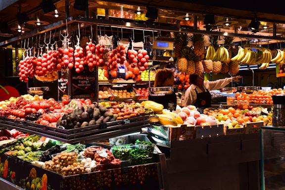 A vibrant market stall filled with a variety of fresh fruits and vegetables. Hanging bunches of bananas and pineapples are on display above stacks of colorful produce including tomatoes, avocados, and papayas. Two people, likely vendors, are engaging with each other behind the counter.