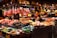 A vibrant market stall filled with a variety of fresh fruits and vegetables. Hanging bunches of bananas and pineapples are on display above stacks of colorful produce including tomatoes, avocados, and papayas. Two people, likely vendors, are engaging with each other behind the counter.