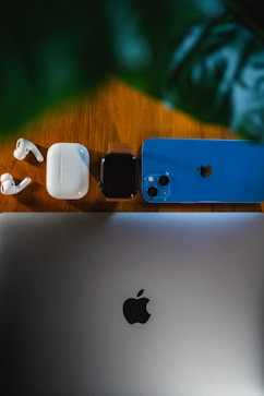 Dispositivos tecnológicos Apple en una mesa de madera: MacBook, iPhone azul, Apple Watch y AirPods