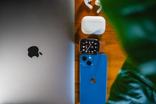 A laptop, smartwatch, smartphone, wireless earbuds, and charging case are neatly arranged on a wooden surface next to a leafy plant. The devices are all from the same brand, featuring a logo of a bitten apple.