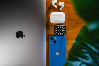 A collection of Apple devices laid on a wooden surface, including a MacBook, AirPods with their case, an Apple Watch, and a blue iPhone. A green leaf partially covers the right side of the image, adding a natural element.