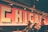 Indoor setting with a large wall mural featuring the word 'CHICAGO' in bold, block letters against a background of red tiles. Multiple people are sitting and engaging in conversations or activities. Lighting fixtures are visible on the ceiling, and a dim, warm ambiance is created by the lighting.