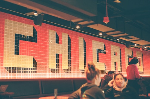 Indoor setting with a large wall mural featuring the word 'CHICAGO' in bold, block letters against a background of red tiles. Multiple people are sitting and engaging in conversations or activities. Lighting fixtures are visible on the ceiling, and a dim, warm ambiance is created by the lighting.
