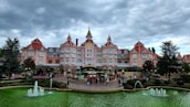 A grand, elaborately decorated pink hotel with multiple pointed towers and a central clock is the main focus. The foreground features a circular garden with vibrant flowers and two fountains spouting water into a vivid green pool. The scene is framed by tall, dark green trees under a moody, overcast sky.