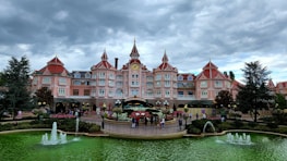 A grand, elaborately decorated pink hotel with multiple pointed towers and a central clock is the main focus. The foreground features a circular garden with vibrant flowers and two fountains spouting water into a vivid green pool. The scene is framed by tall, dark green trees under a moody, overcast sky.