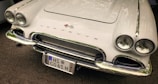 A classic white Corvette with a sleek design, featuring dual round headlights and chrome accents. The car's front grille is prominently displayed with the Corvette emblem above it. The license plate reads HS C061 H.
