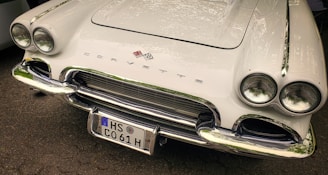 A classic white Corvette with a sleek design, featuring dual round headlights and chrome accents. The car's front grille is prominently displayed with the Corvette emblem above it. The license plate reads HS C061 H.
