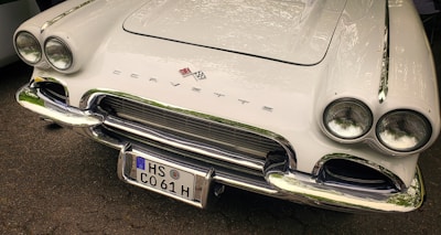 A classic white Corvette with a sleek design, featuring dual round headlights and chrome accents. The car's front grille is prominently displayed with the Corvette emblem above it. The license plate reads HS C061 H.