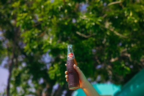 A hand holds up a glass bottle of soda against a backdrop of lush green foliage. The bottle has a red label with white text. Sunlight filters through the leaves, casting a natural, bright atmosphere.