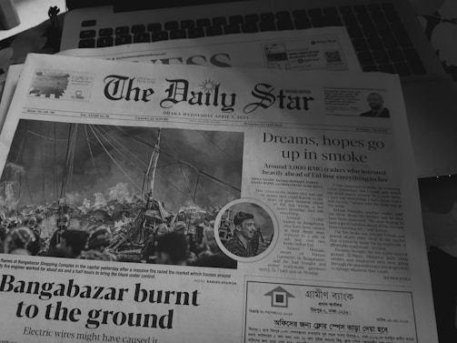 A black and white photo of The Daily Star newspaper with a prominent headline about 'Bangabazar burnt to the ground'. The front page features an image of a large fire with thick smoke and firefighters attempting to control it. Another headline reads 'Dreams, hopes go up in smoke' and discusses the loss due to the fire. The newspaper is placed on top of a laptop keyboard.