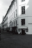 A narrow cobblestone street lined with historical buildings on both sides. The architecture features large windows and traditional design elements. The corner building has a sign reading 'Det Lille Apotek' with small flags displayed. The street has few people and bicycles visible, with an overall quiet and serene atmosphere.
