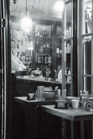 A monochrome scene inside a cozy café or shop. A person stands behind the counter, appearing thoughtful, with shelves of various products behind them. Overhead, warm lighting fixtures hang, creating a soft glow. The counter displays different items, with a container of utensils and cups placed on a nearby table.