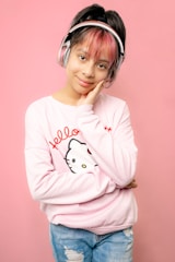 A young person poses against a pink background, wearing headphones with a light pink sweater featuring a Hello Kitty design and blue jeans. Their hair is styled with a touch of pink, and they rest their head on their hand while smiling gently.