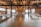 A spacious interior of a rustic event venue with exposed brick walls and wooden beams under a ceiling with string lights. Large wooden tables are arranged in a U-shape, decorated with greenery and place settings for a gathering or celebration.