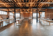 A spacious interior of a rustic event venue with exposed brick walls and wooden beams under a ceiling with string lights. Large wooden tables are arranged in a U-shape, decorated with greenery and place settings for a gathering or celebration.