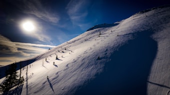 A snowy landscape with a steep mountain slope covered in fresh powder snow. The sun is low in the sky, casting deep shadows across the surface. Several ski tracks curve elegantly down the slope, weaving between sparse trees. The deep blue sky contrasts with the bright white snow, creating a striking visual.