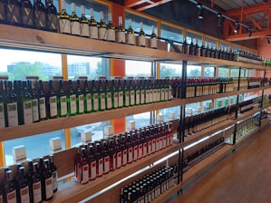 Shelves filled with various bottles of wine arranged in rows, displaying different labels and colors. The setting is an indoor store with large windows allowing natural light to illuminate the bottles from behind.