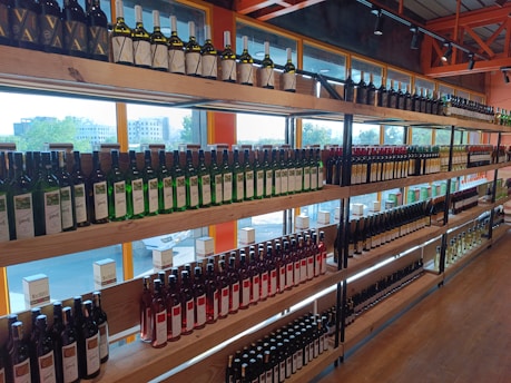 Shelves filled with various bottles of wine arranged in rows, displaying different labels and colors. The setting is an indoor store with large windows allowing natural light to illuminate the bottles from behind.