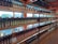 Shelves filled with various bottles of wine arranged in rows, displaying different labels and colors. The setting is an indoor store with large windows allowing natural light to illuminate the bottles from behind.