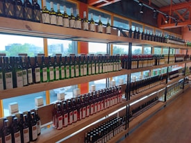 Shelves filled with various bottles of wine arranged in rows, displaying different labels and colors. The setting is an indoor store with large windows allowing natural light to illuminate the bottles from behind.