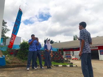 A group of four people, three standing together and one facing them, is outdoors on a concrete area. They are wearing matching blue patterned shirts. One person is taking a photo with a smartphone. A red and blue vertical banner stands beside them, with SMKN written on it. The background includes a low building with a red and gray facade, surrounded by greenery and a cloudy sky.