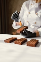 A person dressed in a white chef's jacket is handling rectangular chocolate pastries or cakes. They are wearing black gloves and using a small brush to apply a substance to the pastries. The pastries are arranged neatly on a white surface.