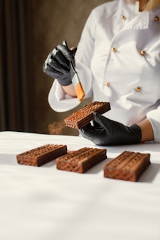 A person dressed in a white chef's jacket is handling rectangular chocolate pastries or cakes. They are wearing black gloves and using a small brush to apply a substance to the pastries. The pastries are arranged neatly on a white surface.
