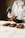 A person dressed in a white chef's jacket is handling rectangular chocolate pastries or cakes. They are wearing black gloves and using a small brush to apply a substance to the pastries. The pastries are arranged neatly on a white surface.