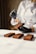 A person dressed in a white chef's jacket is handling rectangular chocolate pastries or cakes. They are wearing black gloves and using a small brush to apply a substance to the pastries. The pastries are arranged neatly on a white surface.