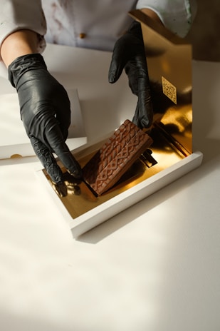 A person wearing black gloves is handling a piece of chocolate with a textured surface, placing it inside a luxurious white and gold box. The setting appears to be clean and well-lit.