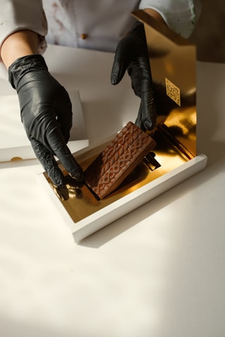 A person wearing black gloves is handling a piece of chocolate with a textured surface, placing it inside a luxurious white and gold box. The setting appears to be clean and well-lit.