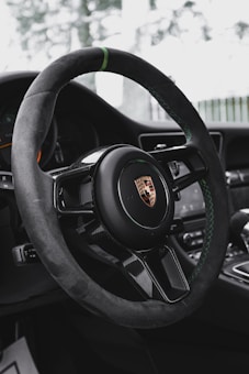 A luxurious car steering wheel with a prominent logo in the center, featuring a sleek design and high-quality materials. The dashboard and part of the interior are visible, hinting at a high-performance vehicle.