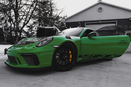 A vibrant green sports car with distinctive black accents is parked on a concrete surface in front of a garage. The car has its driver's side door open, revealing part of the interior. Yellow brake calipers stand out against the black rims. Bare trees and a snow-covered environment can be seen in the background, suggesting a cold weather setting.