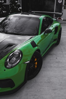 A sleek, high-performance green sports car with a black hood and tinted windows, parked on a concrete driveway outside a building. The car features large black wheels with pronounced yellow brake calipers and a prominent rear wing spoiler. In the background, tall green foliage and a modern building entrance can be observed.