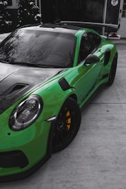 A sleek, high-performance green sports car with a black hood and tinted windows, parked on a concrete driveway outside a building. The car features large black wheels with pronounced yellow brake calipers and a prominent rear wing spoiler. In the background, tall green foliage and a modern building entrance can be observed.