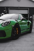 A vibrant green sports car with black accents, having a notable yellow brake caliper, is parked in front of a garage. The building appears to be a workshop or service station with green and white exteriors.