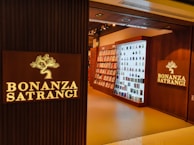 A well-lit retail store entrance features the sign 'Bonanza Satrangi' on a wooden wall. Inside, the store has neatly arranged shelves filled with bottles and boxes, suggesting a display of perfumes or beauty products. The interior is warmly illuminated, enhancing the inviting atmosphere.