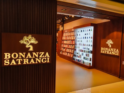 A well-lit retail store entrance features the sign 'Bonanza Satrangi' on a wooden wall. Inside, the store has neatly arranged shelves filled with bottles and boxes, suggesting a display of perfumes or beauty products. The interior is warmly illuminated, enhancing the inviting atmosphere.