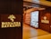 A well-lit retail store entrance features the sign 'Bonanza Satrangi' on a wooden wall. Inside, the store has neatly arranged shelves filled with bottles and boxes, suggesting a display of perfumes or beauty products. The interior is warmly illuminated, enhancing the inviting atmosphere.