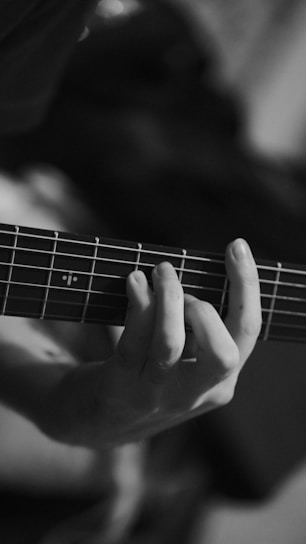 A close-up view of a person's hand playing a guitar, focusing on the fingers pressing down on the strings against the fretboard. The monochrome color scheme gives the image a classic and artistic feel.