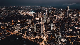 A vibrant city skyline featuring tall skyscrapers illuminated with artificial lights against a dark evening sky. Numerous buildings are closely packed together with roads weaving through the cityscape, showcasing the bustling urban environment.