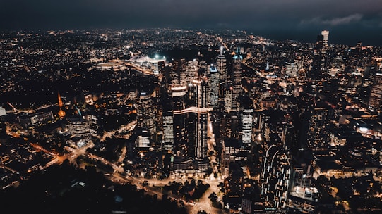 A vibrant city skyline featuring tall skyscrapers illuminated with artificial lights against a dark evening sky. Numerous buildings are closely packed together with roads weaving through the cityscape, showcasing the bustling urban environment.