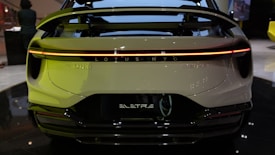 The rear view of an electric vehicle displaying a streamlined design with a prominent spoiler and a continuous LED taillight. The branding 'LOTUS' and 'ELETRE' is visible. The setting appears to be indoors, possibly an auto show or showroom.